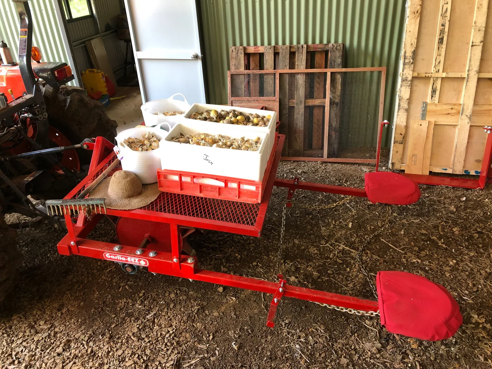 A planter carrying a Garlic-EEZ badge, loaded with trays of seed garlic in a shed and hitched to a tractor, with two cushioned operator seats on the rear rails.